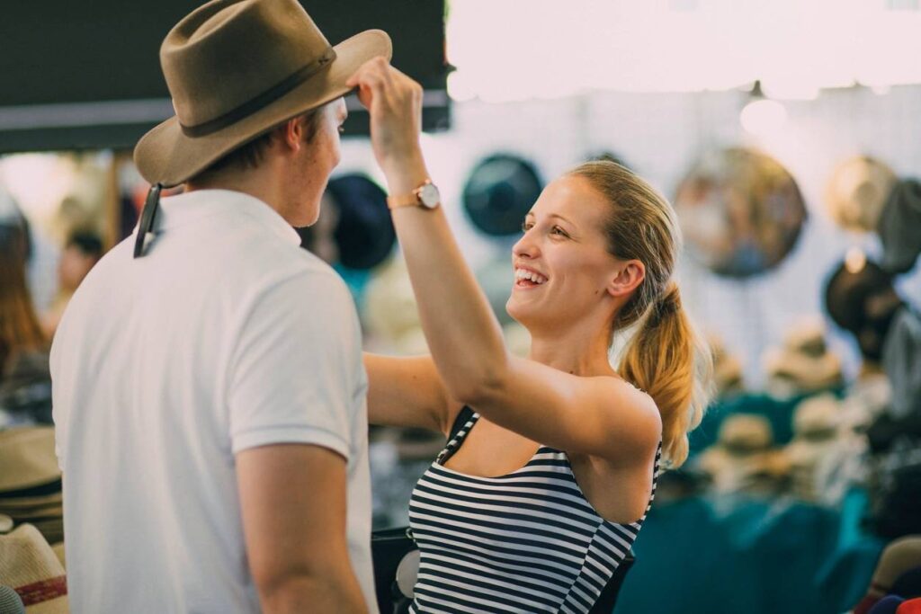Young couple are exploring Queen Victoria Market in Australia. The woman is putting a hat on her boyfriend in a market stall. (Young couple are exploring Queen Victoria Market in Australia. The woman is putting a hat on her boyfriend in a market stall